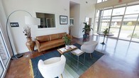 This single-story space makes a strong architectural impression through its dramatic ceiling height and open, airy layout. The living area is anchored by polished concrete floors in a rich, deep tone that add an industrial yet refined character to the interior. Towering floor-to-ceiling windows line one wall, drawing in abundant natural light and offering views of the surrounding exterior landscape, creating a seamless connection between indoors and out.

The seating arrangement is thoughtfully composed, featuring a tan leather sofa paired with modern white accent chairs positioned around a slatted wood coffee table and layered area rugs in cool blue and green tones. A large framed mirror mounted on the white wall enhances the sense of depth and openness throughout the room. Potted tropical plants add organic warmth and color against the crisp white walls.

A transitional archway leads to an adjacent area, suggesting a well-defined flow between spaces. The overall ambiance is contemporary and welcoming, balancing clean architectural lines with warm, curated furnishings to create an inviting and functional environment.