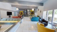 This single-floor space makes a striking impression with its bold, contemporary design and open-plan layout. The interior is anchored by large-format marble-look tile flooring that reflects light beautifully throughout the expansive area, contributing to a bright and airy atmosphere. Recessed lighting is complemented by a pair of decorative globe pendant chandeliers that serve as eye-catching focal points overhead.

The lounge area is furnished with an eclectic mix of colorful seating — including teal, mustard yellow, burnt orange, and charcoal upholstered sofas and armchairs — arranged in multiple conversation groupings that provide both comfort and visual energy. Accent pillows in chevron and geometric patterns add texture and depth to the design.

At the rear of the space, a kitchen and bar counter area is outfitted with bar stools and stainless steel appliances, making it well-suited for entertaining or communal gatherings. Bold yellow accent walls and vibrant artwork frame the kitchen zone, reinforcing the space's lively color palette.

Decorative wall art, including a starburst mirror accent, adds personality to the neutral gray walls. The combination of high ceilings, generous open floor space, and carefully curated furnishings creates an energetic yet inviting ambiance ideal for social interaction and community use.