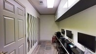 This single-floor space functions as a dedicated computer workstation room, designed to accommodate multiple users in an efficient, organized layout. The room is configured along one wall, where a continuous dark-surfaced built-in desk runs the length of the space, providing ample workspace for multiple monitors and keyboards. Overhead, a row of upper cabinets with dark lower panels and lighter upper doors offers generous storage while keeping the workspace tidy.

The flooring consists of earthy, multi-toned stone-look tiles that add texture and visual warmth to the otherwise neutral interior. The walls are finished in a soft sage green tone, complemented by white crown molding that provides a clean transition to the textured ceiling above. Fluorescent skylight-style fixtures provide consistent, bright overhead illumination throughout the room.

A notable design feature is the decorative partition wall along the opposite side, composed of frosted panels adorned with flowing wave patterns, adding a stylish aesthetic element while subtly dividing the space. A standard interior door provides access deeper into the facility. The overall ambiance is functional and professional, suited for shared or public computing use.