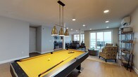 This single-floor space is designed with entertainment and comfort in mind, offering an open-concept layout that seamlessly connects multiple functional areas. The centerpiece of the main room is a full-sized pool table with a bold yellow felt surface, illuminated by a striking trio of brass pendant lights that hang from the ceiling, adding an industrial-chic aesthetic to the space.

The open layout flows naturally into a lounge area furnished with dark leather seating and accent chairs, creating a relaxed atmosphere for gathering. Recessed lighting throughout the ceiling provides consistent, even illumination across the space. Large sliding glass doors or floor-to-ceiling windows line the far wall, flooding the interior with natural light and offering sweeping outdoor views, suggesting a waterfront or elevated setting.

Toward the rear, a compact kitchen is visible, equipped with stainless steel appliances including a range and microwave, accessible via an open pass-through. The flooring throughout is a warm-toned hardwood, contributing to a cohesive, polished look. The neutral gray walls complement the varied furnishings, creating a versatile and well-appointed entertainment environment.