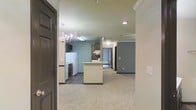 This single-floor residence offers a welcoming open-concept layout that seamlessly connects the kitchen, dining, and living areas. The kitchen is equipped with white appliances, including a refrigerator and range, and features dark cabinetry paired with a central island that provides additional prep space and serves as a natural dividing point between the cooking and living zones. A range hood is positioned above the cooking area, and open shelving adds functional storage. The dining area is illuminated by an elegant chandelier that adds a touch of sophistication to the space. Soft teal-toned walls run throughout, creating a calm and cohesive atmosphere that contrasts pleasantly with the neutral cream ceilings adorned with crown molding. The living area is finished with plush carpeting, lending a comfortable and cozy feel underfoot, while the kitchen and entryway feature darker hardwood-style flooring. Recessed lighting and additional fixtures provide ample illumination throughout. A hallway visible from the main living space leads to additional rooms, suggesting a functional and well-organized floor plan. The overall ambiance is warm, inviting, and practical, making it a comfortable everyday living environment.