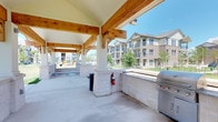 The Grill Area is a thoughtfully designed outdoor amenity space that invites residents to gather, cook, and relax in a comfortable open-air setting. The pavilion is anchored by robust natural wood beams and white structural columns, creating a warm and inviting overhead canopy that provides shade while maintaining an open, airy feel. The flooring is a smooth concrete surface, offering a clean and durable foundation well-suited for outdoor entertaining. A built-in stone counter runs along one side of the space, complementing the stainless steel grill station that is conveniently positioned for easy use. The stone masonry detailing throughout lends a polished, cohesive aesthetic to the overall design. Safety is addressed with a clearly mounted fire extinguisher on one of the support columns. The pavilion opens up to expansive views of the surrounding green landscape, with mature trees and manicured grounds visible in the background. A playground area can be seen nearby, making this an ideal gathering spot for residents of all ages. The multi-story residential buildings in the backdrop provide context for the community setting, reinforcing the grill area's role as a central, social outdoor amenity space.