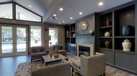 This thoughtfully designed single-floor space exudes a refined, contemporary aesthetic anchored by a striking fireplace framed by a built-in shelving unit finished in deep, muted tones. The shelving provides both decorative and functional storage, displaying curated ceramic vases and woven baskets that add texture and warmth to the room. A sunburst mirror mounted above the fireplace mantel serves as a bold focal point, drawing the eye and complementing the earthy color palette throughout.

The seating arrangement is carefully composed, featuring a mix of plush armchairs and upholstered sofas in neutral tones, centered around a low-profile coffee table adorned with decorative vases. This layout encourages conversation and creates an inviting, communal atmosphere. The flooring transitions between hardwood and a patterned area rug, adding visual interest and defining the lounge zone.

Recessed ceiling lighting provides even, ambient illumination throughout the space, while large windows along one wall flood the interior with natural light and offer views of the surrounding greenery outside. The angled ceiling adds an architectural dimension, contributing to the sense of openness. Overall, the space balances comfort and sophistication, making it an ideal gathering and relaxation area.