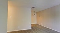 This single-floor space offers a clean, understated aesthetic defined by its neutral color palette and functional layout. The interior is finished with warm beige walls and soft, wall-to-wall carpeting in a muted tone, creating a comfortable and cohesive atmosphere throughout. A textured popcorn ceiling adds a classic character to the space, while standard white baseboards provide a crisp contrast against the flooring. A standout design element is the sleek metallic pendant light fixture, which hangs from the ceiling and serves as a focal point, adding a touch of modern style to the otherwise minimalist interior. The open layout of the living and dining area allows for flexible furniture arrangement and a natural sense of flow between spaces. Ceiling-mounted HVAC vents suggest a central air system, contributing to year-round comfort. The space is bright and evenly lit, with a clean, move-in-ready condition throughout. The overall ambiance is calm and neutral, making it a versatile canvas well-suited to a variety of interior design preferences. This single-story layout provides convenient, accessible living entirely on one level.