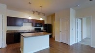 This single-floor space showcases an open-concept kitchen designed for both functionality and style. The kitchen is anchored by a spacious center island topped with granite countertops, providing ample workspace and serving as a natural gathering point. Rich, dark-toned cabinetry lines the kitchen walls, offering generous storage, while black appliances — including a refrigerator, range, and built-in microwave — lend a cohesive, modern aesthetic. A double sink is positioned along the back counter, complemented by additional cabinetry above and below. Overhead, a combination of track lighting and two elegant pendant lights illuminate the workspace effectively. The warm, yellow-toned walls contribute to an inviting ambiance throughout the space. Hardwood-style flooring runs continuously through the kitchen and into the adjoining areas, adding warmth and visual continuity. A paneled interior door is visible beyond the kitchen, hinting at additional rooms within the layout. The open-plan design allows for a natural flow between the kitchen and living areas, making the space feel connected and well-proportioned for everyday living.