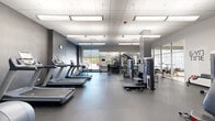 Located at 1807 Glenview Rd in Glenview, Illinois, this well-appointed fitness center spans a single floor and offers a comprehensive workout environment designed to accommodate a wide range of exercise routines. The main exercise room features a dark, rubberized flooring surface and is illuminated by large recessed panel lighting set within a drop-tile ceiling, creating a bright and energizing atmosphere. A row of treadmills lines one side of the room, complemented by elliptical trainers, upright and recumbent bikes, and a full complement of strength-training machines. A dumbbell rack and weight storage area provide additional free-weight options. Full-length mirrors are strategically placed to maximize sight lines throughout the space. Large windows along one wall allow abundant natural light and offer views of the surrounding streetscape, enhancing the open feel of the room. A separate studio area features light-toned flooring, floor-to-ceiling glass partitions, and a wall of mirrors, making it ideal for cycling classes, stretching, or mat-based workouts. This studio also includes a wall-mounted television, yoga mat storage, and additional fitness accessories. Motivational wall graphics and a wall clock add character to the space, reinforcing its purpose-driven, performance-oriented design.