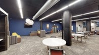 This single-floor space showcases a compelling blend of industrial and contemporary design elements. Exposed metallic ductwork runs along a dark ceiling, complemented by sleek linear LED lighting that illuminates the area efficiently. Striking bold blue accent walls provide a vibrant backdrop throughout, adding energy and character to the environment.

Original structural columns with a dark, textured finish serve as architectural focal points, reinforcing the industrial aesthetic while adding a sense of history to the space. The flooring features patterned commercial carpet in neutral tones, providing visual texture that ties together the various zones within the room.

The layout is thoughtfully divided into distinct functional areas. A lounge zone offers a comfortable sofa and accent chairs arranged around a low table, paired with globe pendant lights and a wall-mounted flat-screen television, creating an inviting setting for informal gatherings. Adjacent collaborative zones feature round and rectangular tables with mixed seating, accommodating group work or small meetings. A whiteboard is also available, supporting brainstorming and presentations. The overall ambiance is open, dynamic, and well-suited to a professional or academic collaborative environment.