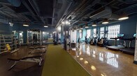 Located in Chicago's Loop at 26 E Adams St, this fitness facility occupies a single floor designed to accommodate a comprehensive range of workout equipment and training zones. The interior features an industrial-inspired aesthetic, with exposed ductwork and conduit running across the dark ceiling, complemented by teal and slate-blue painted walls that give the space a modern, urban character.

The main workout floor is divided into distinct training areas. A dedicated free weights zone includes adjustable benches, dumbbells arranged on tiered racks, and full-length mirrors along the walls to support strength training. A central cable and functional training station anchors the floor, while rows of cardio equipment—including stair climbers, ellipticals, and stationary bikes—line one side of the space. A separate room houses rowing machines and air bikes, surrounded by large windows offering panoramic views of the Chicago skyline.

Flooring transitions between polished concrete, rubberized black mats, and olive-green turf runners, delineating workout zones while providing practical surfaces for varied training. Pendant and track lighting illuminate the space throughout, and wall-mounted mirrors expand the visual depth of the room. A mural-adorned entryway and stairwell access point provide a creative, welcoming transition into the facility.