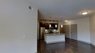 This single-floor residence is designed around an open-concept layout that seamlessly connects the main living area to the kitchen. The kitchen serves as a focal point, showcasing rich dark-toned cabinetry that extends to the upper walls, paired with granite countertops that provide both style and functionality. A central island with a clean white base offers additional workspace and casual seating potential, making it ideal for everyday use or entertaining.

Stainless steel appliances, including a refrigerator, microwave, and range, are integrated neatly into the cabinetry. Pendant lights hang above the island, complementing a track lighting system that directs focused illumination across the cooking and prep areas. The backsplash adds a polished finishing detail to the kitchen design.

Underfoot, dark hardwood flooring runs consistently throughout the space, anchoring the overall aesthetic and adding warmth. The neutral, light-toned walls contrast effectively with the darker finishes, keeping the interior feeling open and balanced. Toward the background, multiple doorways suggest additional rooms beyond the main living and kitchen area. The overall ambiance is modern and functional, with thoughtful design details throughout.