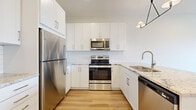 This contemporary single-floor unit offers a clean, modern aesthetic throughout, highlighted by light hardwood-style flooring in the main living areas and plush carpet in the bedrooms. The well-appointed kitchen is a standout feature, showcasing crisp white shaker cabinetry extending to the ceiling, a classic white subway tile backsplash, and stone-look granite countertops. A full suite of stainless steel appliances — including a refrigerator, electric range, over-the-range microwave, and dishwasher — adds a polished, functional touch. A stylish geometric pendant light fixture anchors the kitchen, which opens to the living area via a peninsula with additional counter space. The open-concept living and dining area is bathed in natural light from a large picture window that frames views of the surrounding streetscape. The bedrooms are carpeted for comfort and feature windows that bring in natural light and outdoor views. Closet storage is provided in the bedroom. The bathroom includes a full soaking tub with shower combination, finished in clean white surrounds with wood-look flooring. Bright white walls throughout the unit contribute to an airy, well-lit atmosphere that feels open and inviting.