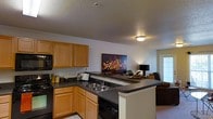 This single-floor residence is designed with an open-concept layout that promotes a sense of connectivity and ease of living. The kitchen is a central highlight, featuring warm honey-toned wood cabinetry that provides ample storage, complemented by dark countertops that offer a striking contrast. A built-in sink and black appliances, including a microwave and dishwasher, contribute to the kitchen's functional design. A peninsula counter extends into the space, creating a natural dividing point between the kitchen and the adjoining living area while also serving as a convenient breakfast bar or serving space. The living area flows directly from the kitchen, creating a cohesive, open atmosphere. Soft ambient lighting from a flush-mount ceiling fixture and a table lamp adds warmth to the space, while a door at the far end hints at access to additional natural light. The textured ceiling adds a subtle architectural detail throughout. The neutral color palette of white walls and light flooring creates a clean, versatile backdrop well-suited to a variety of furnishing styles. Overall, the space balances practicality with a comfortable, inviting ambiance ideal for everyday living and entertaining.
