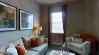 This single-floor space centers around a thoughtfully designed living area that blends comfort with understated style. The room is finished in warm, earthy tones, with taupe-gray walls that create a calm, inviting atmosphere. Natural light filters in through windows dressed with rich burgundy-red curtain panels paired with classic white horizontal blinds, offering both brightness and privacy control. The hardwood-style flooring grounds the space with a clean, contemporary look. Seating is anchored by light gray upholstered sofas accented with a mix of decorative pillows in burnt orange, cream, and patterned tones, adding visual warmth and texture. A glass-topped coffee table with metallic accents introduces a refined touch, while a traditional table lamp with a warm glow contributes to the room's layered lighting. Wall décor includes a large framed abstract artwork in cool and warm mixed tones, serving as a focal point. The overall scale of the living area feels intimate yet functional, well-suited for relaxed everyday living or casual entertaining.