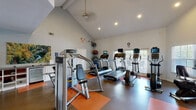 Located in Louisville, KY, this single-story fitness center at Southgate Manor Drive is thoughtfully designed to support a comprehensive workout experience. The space features soaring vaulted ceilings with recessed lighting throughout, creating an open and energizing atmosphere. Large windows fitted with white shutters allow generous natural light to fill the rooms while offering views of the surrounding greenery.

The main workout area is well-appointed with a broad selection of Life Fitness cardio equipment, including multiple treadmills, elliptical trainers, and a recumbent bike, each equipped with individual display screens. Strength training is equally well-represented, with a multi-station cable machine, a full rack of dumbbells ranging across multiple weight increments, adjustable benches, and floor space accented by orange rubber matting. Full-length mirrors in the free weights area enhance the sense of space and assist with form.

A dark granite-topped counter serves as a functional welcome and information station near the entrance. Adjacent to the main gym is a separate lounge and business area featuring warm hardwood-style flooring, a striking arched window, red accent chairs, a bistro table, and a workstation with computer access, providing a comfortable space to relax or work between sessions.