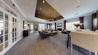 This single-floor community or clubhouse space is designed with a refined, contemporary aesthetic that balances comfort and elegance. The entry foyer sets a welcoming tone with light wood-look tile flooring, soaring ceilings painted in a deep charcoal, and glass-paned French doors that invite natural light and outdoor views.

The main lounge area is expansive and anchored by a striking tray ceiling finished in dark brown, which frames a decorative chandelier with an industrial-modern silhouette. Patterned cut-pile carpeting in neutral tones grounds the space, while large windows dressed in layered drapery flood the room with natural light. A wall-mounted flat-screen television is positioned above a sleek, tile-clad fireplace, flanked by generous sofas and lounge chairs arranged for comfortable gathering.

A dining area along one side features counter-height seating with woven upholstered chairs surrounding a reclaimed-wood-style table, accented by a bold diptych artwork. A credenza with additional display space and wall art complete the lounge periphery. Crown molding throughout adds a polished architectural detail, tying together the space's warm, contemporary design into a cohesive and inviting environment.