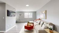 This single-floor unit offers a cohesive, modern aesthetic throughout, unified by soft gray walls, light hardwood-style flooring, and clean white trim. The open living area provides generous space for relaxing and entertaining, centered around a large sectional sofa, a bold terracotta ottoman, and tasteful abstract wall art. Natural light filters in through a white-framed window, complementing the cool, neutral palette. A wall-mounted air conditioning unit ensures year-round comfort. The kitchen is efficiently laid out with crisp white shaker-style cabinetry, a classic white subway tile backsplash, stainless steel appliances including an electric range, and a stainless steel sink — offering a clean and functional cooking environment. The bedroom is a calm and restful retreat, featuring the same light flooring and soft gray walls, accented by a vibrant floral artwork above the bed. A window with an in-unit air conditioner allows for natural light and ventilation. The bathroom is finished with large-format light gray tile flooring, matching wall tile in the tub surround, and a clean white soaking tub and toilet, creating a simple yet polished aesthetic. The overall layout, as seen in the floor plan view, is well-organized and makes efficient use of the available space.