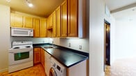 This single-floor unit offers a practical and well-organized layout with distinct living, kitchen, and bathroom areas. The main living space is expansive and open, characterized by warm parquet wood flooring arranged in a classic geometric pattern that adds visual texture throughout. Large windows along one wall frame notable urban views, filling the room with natural light and providing an elevated perspective of the surrounding cityscape.

The kitchen is efficiently designed with warm honey-toned wood cabinetry extending to the ceiling, offering generous storage capacity. Dark countertops provide a striking contrast against the cabinetry and run continuously along an L-shaped configuration, accommodating a stainless steel sink. White appliances include an electric range with an over-the-range microwave. A notable functional feature is the in-unit front-loading washer conveniently integrated beneath the countertop, streamlining laundry within the kitchen footprint. Parquet flooring continues seamlessly into this space.

The bathroom is accessed through a solid wood door and features neutral large-format ceramic tile flooring. It is equipped with a standard toilet and a full shower-tub combination with white tile surround, maintaining a clean and functional aesthetic throughout.
