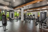 This generously proportioned fitness room offers a full-featured workout environment designed to accommodate a wide range of exercise needs. The space is defined by its striking architectural character, most notably the exposed rustic wood beams that traverse the dark-painted ceiling, lending a warm, lodge-inspired aesthetic to the otherwise contemporary interior. Multiple ceiling fans are strategically positioned throughout to ensure comfortable airflow during workouts.

Durable hardwood-style flooring runs throughout the space, providing a clean and resilient surface suited to both cardio and strength training activities. Large floor-to-ceiling windows line the walls on multiple sides, flooding the interior with natural light and offering views of the surrounding greenery and outdoor balcony areas. A decorative stone wall accent adds further texture and visual interest.

The equipment layout is thoughtfully organized, with a dedicated cardio zone featuring multiple treadmills, elliptical machines, stationary spin bikes, and recumbent bikes. A separate strength training area includes a cable and pulley multi-station machine, a punching bag, weight benches, and additional resistance equipment. A glass-enclosed room at the rear provides a semi-private workout space, enhanced by a vivid landscape mural, and houses additional strength machines. Storage for medicine balls and accessories is also available along the walls.