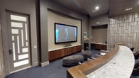 This single-level media lounge is thoughtfully designed to deliver a polished, theater-inspired experience. The focal point of the room is a large wall-mounted flat-screen display set against a curved accent wall finished in a warm neutral tone, creating a cinematic backdrop that draws the eye immediately upon entry. Below the screen, sleek wood-and-stone built-in cabinetry provides ample storage while adding a refined, contemporary aesthetic.

The flooring features deep-toned carpeting that enhances acoustics and contributes to the room's immersive atmosphere. Soft, rounded ottoman seating in a gray upholstery is positioned centrally, offering flexible and casual viewing arrangements. A tiered layout with what appears to be a marble-topped counter at the rear adds a social dimension to the space, ideal for gatherings.

To one side, a dedicated refreshment station — complete with a popcorn machine, compact countertop, and tiled backsplash — adds a fun, functional amenity. Recessed lighting throughout ensures the ambiance remains intimate yet well-illuminated, making this space as practical as it is inviting.