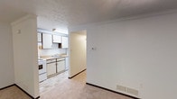 This single-floor residence offers a practical and straightforward layout designed for comfortable everyday living. The kitchen is equipped with white cabinetry, a double-basin sink, and a white electric range, providing ample storage and workspace for meal preparation. Upper and lower cabinets line the kitchen walls, offering generous storage capacity. The kitchen flows naturally into an adjacent hallway and connecting areas, creating an open and accessible feel throughout the unit. Neutral-toned carpeting covers the living area, complementing the light-colored walls and contributing to the space's clean, understated aesthetic. Crown molding accents the ceiling in the living area, adding a subtle architectural detail. A wall-mounted thermostat is conveniently located at the transition between the kitchen and living space. Overhead fluorescent lighting in the kitchen ensures well-lit conditions for cooking and daily tasks, while a floor-level air vent supports efficient climate control. The textured ceiling and consistent white-and-neutral color palette throughout give the space a cohesive and tidy appearance, making it a functional and move-in ready home.