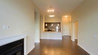 This well-appointed single-floor residence offers a thoughtfully designed open-concept layout that seamlessly connects the living and kitchen areas. Rich, dark hardwood flooring runs throughout the main living space, adding warmth and sophistication to the interior. A classic white fireplace anchors one side of the living area, providing a cozy focal point ideal for relaxing.

The kitchen is a standout feature, highlighted by dark cabinetry contrasted against a light countertop peninsula that doubles as a breakfast bar. Stainless steel appliances — including a refrigerator, microwave, and range — lend a modern, professional feel to the cooking space. Pendant lighting hangs elegantly above the peninsula, complementing the overhead track lighting that illuminates the room.

The open layout allows natural sightlines across the entire space, creating an airy and spacious atmosphere. Neutral-toned walls throughout enhance the sense of openness and provide a versatile backdrop for a variety of décor styles. A hallway visible from the main area leads to additional rooms, suggesting a practical and well-organized floor plan. The overall ambiance is clean, modern, and inviting — making this space both functional and aesthetically refined.