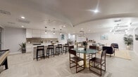 This thoughtfully designed single-floor amenity space offers a seamless blend of social and leisure functions within a clean, modern aesthetic. The layout flows across several distinct zones, each furnished and finished to a high standard.

At the heart of the space is a fully equipped kitchen anchored by a large marble-topped island with bar seating. White flat-panel cabinetry, a tile backsplash, stainless steel appliances, and pendant lighting overhead give the kitchen a polished, functional character. Light herringbone-patterned flooring ties the zones together throughout.

Adjacent to the kitchen, a casual dining area features a round glass-topped table with sculptural gold base seating several guests. Nearby, a billiards table serves as a centerpiece for recreation, complemented by generous lounge seating — including a full sofa, armchairs, and side tables — arranged around a wall-mounted flat-screen television. Contemporary sputnik-style light fixtures add a modern accent overhead.

A standout feature is the intimate banquette nook, finished with arched upholstered panels in soft sage green along the back wall, paired with a warm wood round table and curved accent chairs — creating a cozy, café-like setting.

The space is bathed in recessed lighting throughout, with crisp white walls, curated artwork, and indoor greenery adding warmth and personality to the overall ambiance.