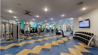 This expansive fitness center is divided into two thoughtfully designed workout zones, each offering a distinct exercise environment. The main gym area features a striking chevron-patterned floor in blue and gold tones, providing a bold visual foundation for a wide array of strength and cardio equipment. This zone is outfitted with cable machines, weight benches, functional training sleds, medicine balls, and a row of treadmills and ellipticals positioned in front of large floor-to-ceiling windows that invite natural light and outdoor views. Ceiling fans and recessed lighting ensure comfort throughout. The walls are adorned with vivid, large-scale geometric murals in teal, navy, gold, and white, featuring bold cityscape and abstract motifs that energize the space. A secondary studio area transitions to warm hardwood flooring and houses a dedicated cycling section with a row of indoor spin bikes, along with full-length mirrors framed in navy, a flat-screen TV, and ample space for group fitness or stretching. This studio benefits from its own large windows, further enhancing the bright and open atmosphere. Glass-panel partitions elegantly separate functional zones while maintaining visual connectivity. Together, both areas form a cohesive, professionally appointed fitness facility designed to support a full range of workout routines.