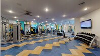 This expansive fitness center is divided into two thoughtfully designed workout zones, each offering a distinct exercise environment. The main gym area features a striking chevron-patterned floor in blue and gold tones, providing a bold visual foundation for a wide array of strength and cardio equipment. This zone is outfitted with cable machines, weight benches, functional training sleds, medicine balls, and a row of treadmills and ellipticals positioned in front of large floor-to-ceiling windows that invite natural light and outdoor views. Ceiling fans and recessed lighting ensure comfort throughout. The walls are adorned with vivid, large-scale geometric murals in teal, navy, gold, and white, featuring bold cityscape and abstract motifs that energize the space. A secondary studio area transitions to warm hardwood flooring and houses a dedicated cycling section with a row of indoor spin bikes, along with full-length mirrors framed in navy, a flat-screen TV, and ample space for group fitness or stretching. This studio benefits from its own large windows, further enhancing the bright and open atmosphere. Glass-panel partitions elegantly separate functional zones while maintaining visual connectivity. Together, both areas form a cohesive, professionally appointed fitness facility designed to support a full range of workout routines.