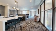 Located at 6405 Rex Ln in Alpharetta, GA, this single-floor communal space is thoughtfully designed to accommodate both collaborative work and relaxed social interaction. The open layout is anchored by a striking herringbone-patterned carpet in neutral tones that adds texture and visual depth throughout the room.

A central conferencing area features a sleek white table framed by bold royal blue upholstered booth seating, creating a polished and functional focal point. Adjacent to this, a dedicated workstation zone is outfitted with a long white work surface, ergonomic black office chairs, and semi-private partitioned cubicles ideal for focused tasks. A modern globe pendant light fixture with brass accents hangs overhead, lending warmth and contemporary style to the work area.

On the opposite end, a comfortable lounge cluster of curved, light-toned armchairs invites casual conversation beneath curated abstract wall art. Floor-to-ceiling black-framed glass panels and windows frame outdoor views of the surrounding courtyard and pool area, flooding the space with natural light and maintaining a bright, open atmosphere. Recessed ceiling lighting supplements the natural illumination throughout. The overall design strikes a balance between professional functionality and inviting comfort, making it a versatile and well-appointed community space.