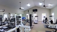 Located at 4223 I-20 Frontage Road in Vicksburg, Mississippi, this single-floor fitness center is designed to support a well-rounded workout experience. The space features soaring vaulted ceilings that lend an open, airy atmosphere, complemented by ceiling fans that help maintain comfortable airflow throughout the room. Multiple windows allow generous natural light to fill the space, creating a bright and energizing environment.

The facility is outfitted with a comprehensive selection of exercise equipment. Strength training options include a multi-station cable machine and a free weights rack with dumbbells, while stability and flexibility training are supported by exercise balls and floor mats. For cardiovascular workouts, the space provides treadmills and an elliptical machine, ensuring a variety of aerobic options for users.

A large wall mirror runs along one side of the room, enhancing the sense of space and allowing members to monitor their form during workouts. A wall-mounted flat-screen television adds an entertainment element to the fitness experience. The neutral-toned walls and light carpeted flooring contribute to a clean, welcoming aesthetic. Overall, this fitness center offers a well-equipped, comfortable, and functional space for a diverse range of exercise routines.