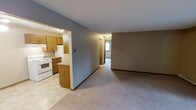 This single-floor residence offers a straightforward and functional layout designed for comfortable everyday living. The kitchen is positioned off the main living area, separated by a half-wall that creates an open, connected feel while maintaining a defined cooking space. It features warm oak cabinetry, white appliances including a range and refrigerator, and light-toned flooring that contrasts nicely with the rest of the unit. The adjoining living area is generously proportioned, finished with neutral wall-to-wall carpeting and soft gray walls that lend a calm, understated ambiance throughout. A hallway extends from the living space toward additional rooms, offering a glimpse of natural light filtering in from a window at its end, suggesting a bright and accessible private area. Warm wood-toned baseboards and door trim run consistently throughout, providing a cohesive design element that ties the spaces together. The overall atmosphere is clean, practical, and move-in ready, making it well-suited for those seeking a low-maintenance, single-level living arrangement with a functional open floor plan.
