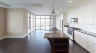 Located on the 17th floor at 11550 Old Georgetown Road in Bethesda, Maryland, this well-appointed residence offers a thoughtfully designed single-level layout with refined finishes throughout. The open-concept kitchen and living area serves as the heart of the home, featuring floor-to-ceiling windows that flood the space with natural light and frame sweeping elevated views of the Bethesda skyline and treetops. The kitchen is equipped with stainless steel appliances, white shaker cabinetry, a white subway tile backsplash, dark granite countertops, and a large central island with a built-in sink and pendant lighting overhead. Rich, dark hardwood flooring extends throughout the main living areas, while a tray ceiling detail adds architectural interest to the living room. The bedrooms are carpeted in neutral tones and feature large windows with views of the surrounding landscape. The bathrooms are finished with striking black-and-white mosaic tile flooring, subway-tiled tub surrounds, and a double-sink vanity with dark cabinetry, granite countertops, and an illuminated mirror. Clean lines, neutral warm tones, and quality materials define the overall aesthetic of this elevated Bethesda residence.
