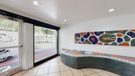 This single-story lobby serves as the welcoming entrance and leasing hub of a residential community with strong University of Florida ties. The space is finished with neutral, large-format ceramic tile flooring accented by decorative mosaic inlays that echo throughout, creating a cohesive and lively aesthetic from entry to elevator bank.

The most striking feature is a custom, organically shaped bench constructed from concrete and adorned with a colorful mosaic tile surface in the University of Florida's signature orange, blue, and green palette. Mounted directly above on the white wall is a large mosaic panel boldly displaying the Florida Gators logo, reinforcing the community's collegiate identity. Recessed lighting throughout keeps the space well-illuminated and clean.

Large black-framed storefront windows and glass entry doors line the street-facing wall, flooding the lobby with natural light and providing open views to the surrounding streetscape. A dedicated leasing office window is accessible from within the lobby.

Toward the rear, a pair of stainless steel elevators provides vertical access to upper floors, flanked by informational signage. The overall ambiance is energetic, branded, and welcoming—well-suited to a student-oriented residential community near campus.