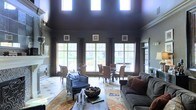 This single-floor space makes a striking impression with its dramatically high ceilings finished in a deep, dark hue, creating a bold architectural contrast against the lighter walls adorned with classic crown molding. Three oversized multi-pane windows dominate the rear wall, flooding the interior with natural light and offering views of the surrounding outdoor greenery. Above, a row of clerestory windows further enhances the brightness, drawing the eye upward and emphasizing the generous vertical scale of the room.

The open-plan layout seamlessly integrates a living area and a dining space. The living zone features a plush gray sofa accented with warm rust and brown throw pillows, paired with a navy blue accent chair and a low white coffee table resting atop a traditional patterned area rug. Decorative framed wall art and a fireplace mantle add refined touches to the walls.

The dining area, positioned near the windows, accommodates a round table surrounded by upholstered chairs, creating an inviting setting for gatherings. The light-toned tile flooring ties both zones together cohesively. Overall, the space exudes a sophisticated, moody elegance balanced with warmth and natural light.