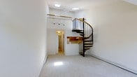 Located at 1202 Crescent Dr in Tarrytown, NY, this two-bedroom, one-bathroom loft offers a thoughtfully designed two-story layout that maximizes both space and character. The main living area is bright and open, anchored by a striking spiral staircase with a warm wood handrail and sleek black metal balusters — a standout architectural feature that serves as both a functional element and a visual centerpiece. Plush carpeting extends throughout the living space, adding warmth and comfort underfoot. Overhead, skylights flood the interior with abundant natural light, enhancing the airy, open atmosphere. The upper loft level, accessible via the spiral staircase, is enclosed by a wood-railed balcony that overlooks the main living area below, creating an engaging sense of vertical space. The kitchen features wood-toned cabinetry and is conveniently positioned adjacent to the main living area, visible through a pass-through opening. Track lighting in the living space and a ceiling fan provide practical illumination and climate comfort. Baseboard heating ensures year-round warmth. The overall ambiance is clean, open, and inviting, with white walls and natural light creating a bright, welcoming environment throughout this distinctive loft-style residence in Tarrytown.