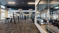 This expansive fitness center is designed to accommodate a comprehensive range of workout needs within a thoughtfully organized, single-level layout. The space is defined by soaring double-height ceilings in key areas, floor-to-ceiling windows that flood the interior with natural light, and an exterior view that adds an open, airy quality to the environment.

The main strength training area features patterned, multi-toned carpet flooring and is well-equipped with a full rack of dumbbells, weight benches, cable machines, and various selectorized strength machines. Large wall mirrors extend across multiple sections, enhancing the sense of space. Linear pendant and recessed lighting illuminate the area against a dark, dramatic ceiling.

A dedicated cardio zone houses stair climbers and other cardio machines, positioned beneath tall windows that provide natural light and outdoor views. Adjacent to this is a glass-enclosed cycling studio with wood-style flooring, stationary bikes, step platforms, and direct access to an outdoor area.

A separate glass-partitioned room serves as a flex or training space, furnished with a wall-mounted TV, storage cubbies, and seating. A water fountain and additional storage cabinetry are also present, rounding out the practical amenities throughout the facility.