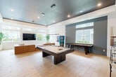 Located at 6136 Blanding Boulevard in Jacksonville, Florida, this single-floor community lounge space offers a well-appointed and thoughtfully designed environment across two distinct living areas. The layout provides a generous, open feel enhanced by soaring ceilings and an abundance of natural light streaming through large windows and clerestory transoms that frame views of the surrounding outdoor spaces.

The first living area serves as a relaxed entertainment hub, anchored by a plush cognac leather sectional sofa and a wall-mounted flat-screen television. Light wood-look flooring runs throughout, complementing the crisp white walls and a striking dark tray ceiling fitted with an elegant circular pendant chandelier. Lush potted palms add warmth and a tropical touch, while a wall-mounted game board provides an interactive focal point. A pool table is also visible, adding a recreational dimension to this gathering space.

The second living area takes on a more intimate lounge character, featuring soft taupe armchairs arranged around low white coffee tables atop a textured area rug. A large-scale diptych wall art depicting palm trees against a pastel sky serves as a bold decorative centerpiece, reinforcing the space's coastal aesthetic. Together, both areas create a cohesive, modern atmosphere ideal for socializing and relaxation.