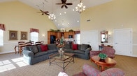 Located at 105 Amberley Dr in Blue Bell, Pennsylvania, this single-floor community space offers an inviting and well-appointed environment designed for gathering and socializing. The most striking architectural feature is the dramatic vaulted ceiling, which rises to impressive heights and is adorned with multiple ceiling fans and elegant chandelier lighting, creating a bright and airy atmosphere throughout.

The open-concept layout seamlessly connects a generously sized living area with a fully equipped kitchen. The living space is furnished with plush, dark upholstered sofas and accent chairs accented with rich red throw pillows, arranged around a glass-top coffee table — providing a warm and comfortable setting for residents or guests. Decorative red-checkered valances frame the windows, adding a charming, classic touch to the interior.

The kitchen area features warm wood cabinetry extending to the upper walls, black appliances, and ample counter space, making it well-suited for catering and events. Recessed lighting and pendant fixtures supplement the natural light filtering in through multiple windows. The neutral-toned flooring ties the space together, lending a cohesive and polished look. Overall, the space exudes a welcoming, community-oriented ambiance ideal for a variety of social functions.