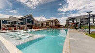This expansive outdoor amenity space centers around a large, resort-style swimming pool with crystal-clear blue water and integrated in-pool lounge shelves, ideal for leisurely relaxation. The pool deck is generously sized, accommodating numerous chaise lounges, red Adirondack chairs, and patio umbrellas arranged to maximize sunbathing and socializing opportunities. A prominent clubhouse anchors one end of the pool area, featuring a bold blue board-and-batten exterior with large windows and stone accent details, lending the space a modern yet welcoming architectural character. Adjacent to the pool, a wood pergola-covered lounge area provides a shaded retreat outfitted with vibrant yellow wicker chairs, a fire pit table, and bar-height seating, creating an inviting gathering spot. A wrought iron fence borders the perimeter, offering a sense of enclosure while maintaining open sightlines to the surrounding green lawn and multi-story residential buildings beyond. String lights are strung overhead in select areas, suggesting an inviting atmosphere during evening hours. The overall design blends contemporary residential architecture with active outdoor living, offering a well-appointed communal space suited for warm-weather enjoyment and community engagement.