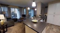 This single-story space showcases an open-concept layout that integrates the kitchen and living area into one cohesive, functional environment. The kitchen is anchored by a large central island with a dark wood-tone laminate countertop, housing a deep double-basin stainless steel sink with a contemporary arc faucet. Pendant lighting hangs above the island, providing warm, focused illumination that complements the overall ambiance. Cabinetry in a neutral tone lines the kitchen perimeter, offering ample storage.

The living area flows naturally from the kitchen, featuring plush carpeted flooring and a comfortable seating arrangement centered around a round wooden coffee table. A flat-screen television is positioned against the wall, making the space well-suited for both relaxing and entertaining. Patterned drapery in a geometric print frames the large windows and sliding glass doors, which allow generous natural light to fill the interior while providing direct access to an outdoor patio area. The warm, neutral color palette throughout creates a cohesive and inviting atmosphere across the entire open floor plan.