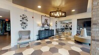 This single-floor community clubhouse is designed with a cohesive modern aesthetic, anchored throughout by a striking multi-toned hexagonal tile floor that flows seamlessly from room to room. The welcoming entrance lobby greets visitors with a geometric chandelier, curated wall art, a decorative credenza, and comfortable seating, setting an inviting tone for the entire space.

The open-plan layout connects the lobby to a fully equipped kitchen featuring dark cabinetry, marble-style countertops, stainless steel appliances, and an island with bar seating — ideal for social gatherings. Adjacent to the kitchen, the game room offers dedicated areas for billiards and shuffleboard, accented by large-scale artwork and recessed lighting with a tray ceiling detail.

A business center provides a built-in workstation with upper cabinetry and marble countertops, offering a quiet, functional workspace. Nearby, a cozy casual dining nook with high-top seating benefits from floor-to-ceiling windows that frame outdoor greenery and fill the area with natural light.

The professional office suite includes a spacious manager's office with large windows overlooking the exterior, a private meeting room with upholstered seating, and a dedicated staff area with a glass-paneled entryway. Throughout, the space balances contemporary design with practical amenities in a polished, well-appointed environment.