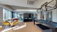 This sophisticated amenity space is defined by its contemporary design and diverse array of social and recreational areas, all connected across a single expansive floor. The grand lobby serves as the centerpiece, boasting soaring double-height ceilings, a striking sculptural wall accent, a sleek reception desk adorned with hexagonal wood detailing, and a cascading pendant light fixture. Natural light pours in through floor-to-ceiling windows, offering views of a well-appointed outdoor courtyard and pool area. Comfortable seating arrangements with modern furnishings and plush area rugs create inviting lounge zones throughout. A dedicated entertainment lounge features a large sectional sofa, teal accent chairs, and a refined fireplace set against a dark stone surround. Adjacent to this is a billiards area with a professional pool table and a stylish geometric pendant light overhead. The private screening room impresses with rows of plush leather reclining theater seats, a deep navy tray ceiling with recessed lighting, and a large wall-mounted display screen. A separate game room offers round card tables surrounded by vibrant yellow chairs in a bright, window-lit setting. The space is finished throughout with warm hardwood flooring and thoughtful, curated décor that ties the entire experience together.