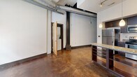 Located at 101 E Sixth St in downtown Winston-Salem, NC, this unit showcases a distinctive industrial aesthetic throughout. The open living area features polished concrete floors, soaring ceilings with exposed ductwork, and structural columns that reflect the building's warehouse heritage. A kitchen peninsula with granite countertops and dark wood cabinetry anchors the main living space, complemented by pendant lighting. The bedroom offers a spacious closet and large factory-style windows with views of the surrounding streetscape. A hallway provides access to an in-unit stacked washer/dryer. The bathroom is finished with tile flooring, a dark wood vanity, granite countertop, and a full-width mirror.