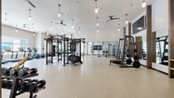This expansive fitness center is thoughtfully designed across a single floor, offering a comprehensive workout environment that caters to a wide range of exercise disciplines. The space is divided into two distinct zones: a large open main gym area and a separate studio room.

The main gym area features soaring ceilings accented by rustic wood columns that add warmth to the modern aesthetic. Durable speckled rubber flooring provides a safe, high-performance surface throughout. The space is equipped with a functional trainer rig, Smith machine, free weight stations with a full dumbbell rack, cable machines, and a punching bag. A row of treadmills, ellipticals, and other cardio machines lines the perimeter beneath large windows that allow abundant natural light. Full-length mirrored walls amplify the sense of space and aid in form monitoring. Pendant lighting fixtures and ceiling fans maintain a bright, well-ventilated atmosphere, while wall-mounted flat-screen TVs offer entertainment during workouts.

The adjoining studio room features light hardwood flooring, a tray ceiling with recessed lighting, and large frosted windows. It accommodates spin bikes, a medicine ball rack, and additional cardio equipment, making it ideal for group or individual training sessions. The overall design strikes a balance between modern functionality and inviting warmth.
