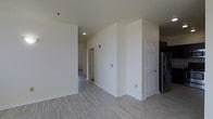 This single-floor space offers a fresh, modern aesthetic characterized by crisp white walls and light wood-style plank flooring that runs throughout the main living area, creating a bright and cohesive feel. The open layout allows for easy flow between the main living space, a connecting hallway, and the adjacent kitchen area. A stainless steel refrigerator is partially visible near the kitchen entry, hinting at contemporary appliances within. Interior doors feature classic six-panel designs painted white, complementing the clean, neutral color palette throughout. Recessed and ceiling-mounted lighting ensures the space remains well-lit, while electrical outlets are conveniently placed along the walls. The hallway visible in the background leads to additional rooms, suggesting a functional and organized floor plan. The overall ambiance is clean, minimalist, and move-in ready, with neutral finishes that provide a versatile backdrop for a variety of interior styles. The uncluttered design and consistent flooring throughout contribute to a sense of openness and ease of movement across the space.