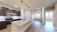 This well-appointed single-floor residence offers a thoughtfully designed open-concept layout that seamlessly connects the kitchen, dining, and living areas. The kitchen serves as the centerpiece of the home, showcasing rich dark cabinetry that reaches toward the ceiling, a striking herringbone-patterned tile backsplash, and stainless steel appliances including a refrigerator, range, and microwave. A generous central island topped with light speckled granite countertops provides both additional prep space and a natural gathering point, elegantly illuminated by a row of glass pendant lights. Warm wood-style flooring flows continuously throughout the entire space, lending a cohesive and inviting feel. The open living area benefits from multiple windows dressed with blinds, allowing natural light to fill the room while a ceiling fan adds comfort. Bedrooms are well-sized with ample natural light and easy access to bathrooms featuring granite-topped vanities with dark wood cabinetry and subway-tiled soaking tubs with brushed nickel grab bars. Walk-in closets with built-in shelving and hanging rods provide generous storage. Neutral warm-toned walls and clean white trim throughout the home create a bright, modern, and move-in-ready atmosphere.