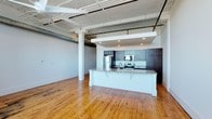Unit 602 is a 1-bedroom, 1-bathroom residence that showcases a striking industrial-loft aesthetic. The open-concept layout is anchored by soaring ceilings with exposed ductwork, structural beams, and white-painted trusses that lend a distinctive character to the space. Warm-toned hardwood floors run throughout, adding a natural richness that complements the industrial overhead elements.

The kitchen is centrally positioned and designed for both function and style. It features a large island with a granite countertop and an undermount sink, providing ample workspace and a natural gathering point. Dark cabinetry lines the walls, offering generous storage, while stainless steel appliances — including a refrigerator, range, and built-in microwave — complete the modern setup.

Recessed lighting under a dropped soffit illuminates the kitchen area, creating a well-lit cooking zone that contrasts with the expansive open ceiling of the living area. Structural columns punctuate the space, reinforcing the loft's architectural heritage. The overall ambiance blends raw industrial character with refined finishes, resulting in a unit that feels both spacious and thoughtfully designed.