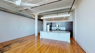 Unit 602 is a 1-bedroom, 1-bathroom residence that showcases a striking industrial-loft aesthetic. The open-concept layout is anchored by soaring ceilings with exposed ductwork, structural beams, and white-painted trusses that lend a distinctive character to the space. Warm-toned hardwood floors run throughout, adding a natural richness that complements the industrial overhead elements.

The kitchen is centrally positioned and designed for both function and style. It features a large island with a granite countertop and an undermount sink, providing ample workspace and a natural gathering point. Dark cabinetry lines the walls, offering generous storage, while stainless steel appliances — including a refrigerator, range, and built-in microwave — complete the modern setup.

Recessed lighting under a dropped soffit illuminates the kitchen area, creating a well-lit cooking zone that contrasts with the expansive open ceiling of the living area. Structural columns punctuate the space, reinforcing the loft's architectural heritage. The overall ambiance blends raw industrial character with refined finishes, resulting in a unit that feels both spacious and thoughtfully designed.