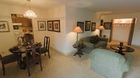 This single-floor space features an open layout that seamlessly connects the living and dining areas, creating a cohesive and functional flow. The living area is anchored by a plush, sage-green sofa accented with decorative pillows, complemented by a classic round pedestal side table that adds a traditional touch. Warm ambient lighting from table lamps with soft shades creates an inviting atmosphere throughout the space. The carpeted flooring in a neutral tone runs throughout, adding comfort underfoot and contributing to the overall warmth of the interior. A collection of framed artwork is thoughtfully arranged along the walls, adding visual interest and personality to the space. The dining area, partially visible to the left, features traditional dark wood chairs and a chandelier-style light fixture overhead, reinforcing the classic aesthetic. A partial wall divides the two areas while maintaining an open feel. Built-in shelving is glimpsed in the background, offering practical storage. The overall ambiance is cozy and well-appointed, with warm tones and traditional furnishings creating a comfortable, lived-in atmosphere.