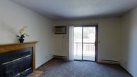 This single-floor space presents a practical and comfortable layout designed for everyday living. The main living area is carpeted and features a classic fireplace with a wood-trimmed mantel and tile surround, serving as a natural focal point of the room. A sliding glass door floods the space with natural light and opens directly onto a private balcony, offering a pleasant outdoor view with glimpses of surrounding trees. A wall-mounted air conditioning unit provides climate control, complemented by baseboard heating along the perimeter walls for year-round comfort. The textured ceiling adds a traditional character to the interior. The neutral wall tones and understated finishes create a clean, versatile backdrop well-suited to a variety of furnishing styles. Overall, the space balances functional amenities with a relaxed, approachable ambiance that makes it easy to personalize and settle into.