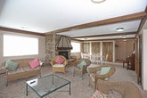 This single-story home showcases a classic, traditional design highlighted by rich dark wood beam ceilings that extend throughout the main living areas, adding warmth and architectural character. The expansive living room serves as the heart of the home, anchored by a striking floor-to-ceiling stone fireplace with a distinctive hood — an eye-catching focal point that complements the casual wicker seating arrangement. Multiple windows along the walls invite generous natural light, creating a bright and welcoming atmosphere. Adjacent to the living room, a dining area accommodates a full dining set and features an elegant chandelier overhead, with a sliding glass door offering direct access to an outdoor deck or balcony with views of surrounding greenery. A secondary dining nook near the kitchen provides additional flexible dining space. The kitchen is appointed with rich, dark wood cabinetry and modern appliances, connected to the main living areas through warm wood-trimmed doorways. Throughout the home, soft carpeting and textured ceilings reinforce a cozy, lived-in character. A dedicated alcove houses an upright piano, hinting at the space's versatility. An adjoining room lined with bookshelves adds a charming library nook, completing this well-appointed and thoughtfully designed home.